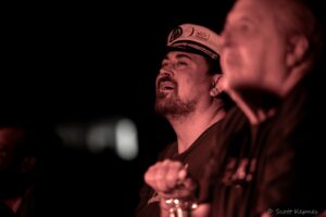 Bearded man wearing a captain's hat holding a drink at a dimly lit party, red lighting in the background.