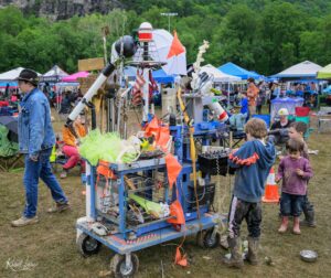 DIY art sculpture built from random metal and household items on a wheeled cart at an outdoor festival with tents in the background.