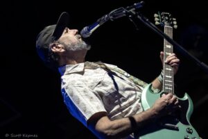 Male guitarist sings into a microphone on stage, wearing a cap and floral shirt with a mint-green electric guitar
