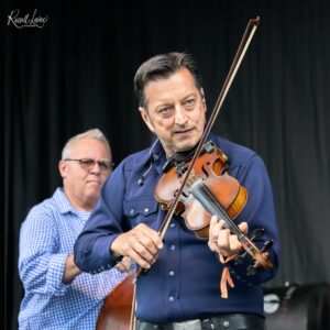 Violinist in a dark blue shirt playing a violin on stage, a second musician in a checkered shirt visible behind him against a black curtain backdrop, with a Russell Leine photographer logo in the top left.