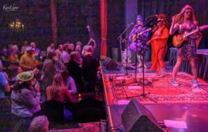 Live band on a brightly lit stage performing while a cheering audience watches from the front barrier in a dim, colorful venue.