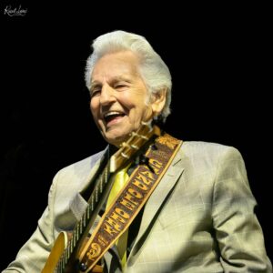 Close-up of smiling elderly man with white hair, wearing a light plaid suit and guitar strap on a dark stage area.