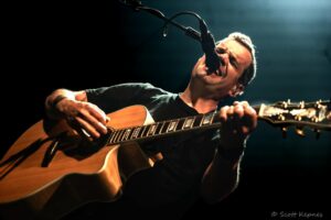 Male musician singing into a microphone while playing an acoustic guitar on stage.