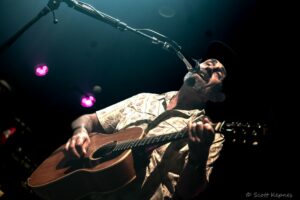 Male musician on stage singing into a microphone while playing an acoustic guitar, wearing a hat under dark blue stage lights.