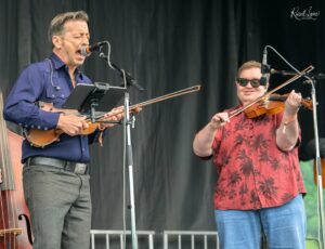 Two male musicians perform on a stage: one sings into a microphone while playing a violin, the other plays a violin beside him, wearing sunglasses and a red patterned shirt.
