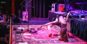 Performer in a black outfit bends backward on a red patterned rug, holding a microphone during a dimly lit live concert. Audience claps in the background.