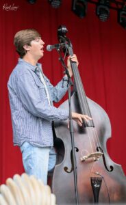 Male musician singing into a microphone while playing a large double bass on a stage with a red curtain backdrop.
