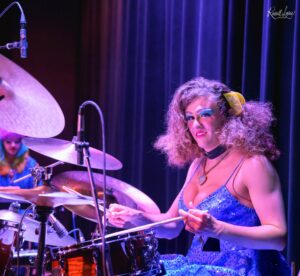 Female drummer in a blue sequined dress with blue makeup playing drums on a dimly lit stage with purple lighting and cymbals visible.