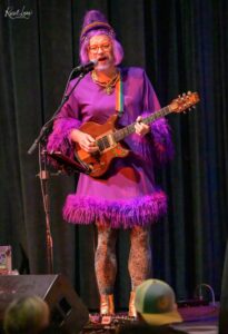 Performer in a purple feather-trim dress with purple wig, red glasses, and rainbow guitar strap singing into a microphone while playing an electric guitar on stage.