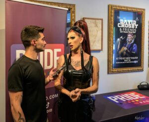 Drag queen in black latex outfit being interviewed by a man holding a microphone, indoors by event posters on the wall.