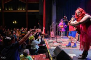 Live stage performance with performers in colorful outfits playing guitars while the cheering audience claps and reaches toward the stage.