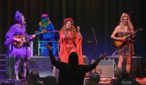Live concert scene with four performers on stage: a purple-clad guitarist left, rainbow-haired keyboardist center, red-haired singer in orange dress center, and blonde guitarist right; audience hands raised in front.