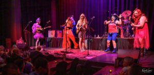 Group of diverse musicians performing on a colorful stage with guitars and a drummer, audience watching.