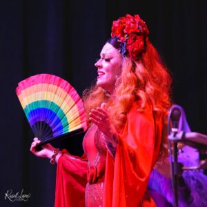 Performer in a red costume with a large red flower headpiece, smiling while holding a rainbow fan on stage.