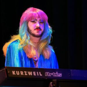Drag performer with long rainbow hair and dramatic makeup on a dark stage, wearing a blue outfit under vibrant lighting.