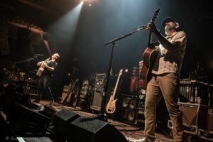 Two musicians perform on a dimly lit stage, the guitarist on the right wearing a cap and patterned shirt while playing an acoustic guitar and singing into a microphone. A second guitarist stands left with a guitar, surrounded by amps and stage gear in a concert setting.