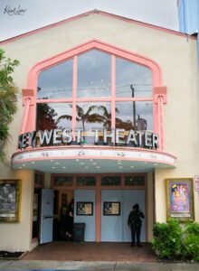 Front entrance of the Key West Theater with a pink arched window frame, glass doors, a marquee reading 'KEY WEST THEATER', and a security guard nearby.