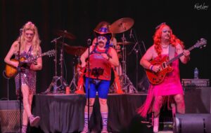 Three performers on a dark stage: left guitarist in a metallic dress, center singer in a red mesh top with a patriotic hat, right guitarist in a pink fringe dress; drums behind them.