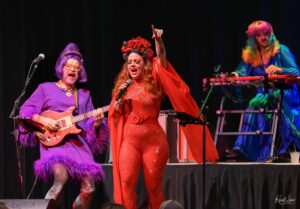 Vibrant stage performance with three singers: a red-dressed lead with flower headpiece centers stage, a purple-clad guitarist left, and a rainbow-haired keyboardist right.