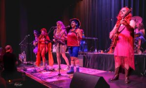 A group of performers on a colorful stage, singing into microphones with instruments, under pink and purple stage lights. The lead singer wears a red outfit and blue hair, while others wear bright costumes and accessories.