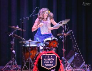 Female drummer on stage, wearing a blue outfit, playing a drum kit with a sign that reads "IT'S A RAINBOW FULL OF SOUND" in front and a red feather boa. behind black stage curtains.
