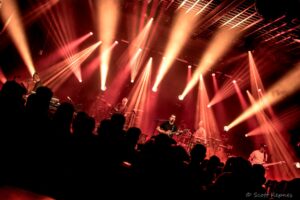 Band performing on stage under red and orange spotlights with a silhouetted crowd.
