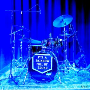 Blue-lit image of a drum kit on a stage with cymbals, microphones, and stands; bass drum front reads 'It's a rainbow full of sound.'