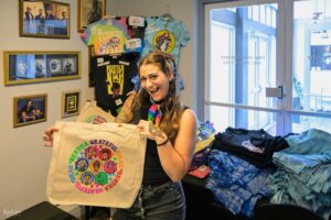 Smiling woman holds a beige tote bag with a colorful circular graphic, standing in a room with tie-dye shirts on a rack behind her.