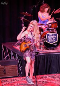 Female musician on stage, singing into a microphone while playing an orange electric guitar; drummer in blue behind her under pink stage lights, banner reads 'It's a Rainbow Full of Sound'