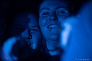 Two friends close together, smiling in blue-tinted low light at a social event.