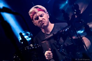 Drummer with blond hair and red bandana playing drums on a blue-lit stage, concentrating on the beat.