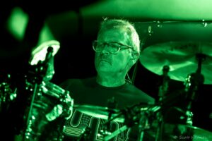 Close-up of a man wearing glasses playing drums on a green-lit stage.