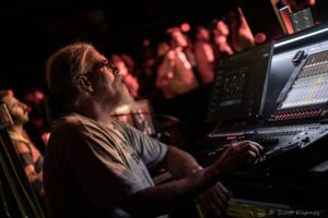 Close-up of an older man with glasses and gray beard operating a large audio mixing console at a concert scene. Reddish stage lighting and blurred crowd in the background.