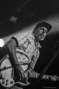 Male guitarist wearing a patterned shirt and hat, playing an electric guitar on stage under a spotlight (black and white)