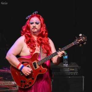Drag queen with a long red wig and dramatic makeup playing a red electric guitar on a dark stage background