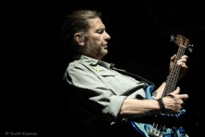 Side view of a man playing a blue bass guitar on a dark stage, wearing in-ear monitors.