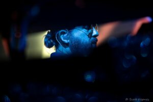 Profile of a bearded man with glasses in blue stage lighting, wearing an in-ear monitor.
