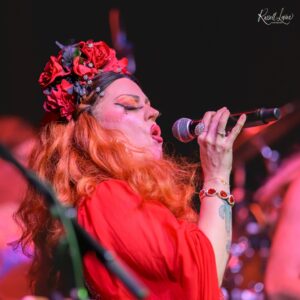 Singer wearing a red flower crown and red dress, singing into a microphone on stage with eyes closed.