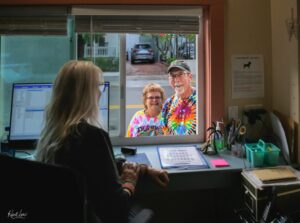 Two people in colorful tie-dye shirts smile at a service window from outside, while a receptionist sits inside with computers and office supplies nearby.