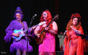 Three drag performers on a dark stage playing guitars and singing, bathed in colorful stage lighting (purple, pink, red).