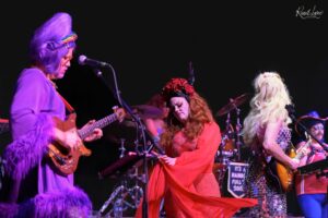 A vibrant stage performance with three women: purple-haired guitarist on left, red-dressed singer center, and blonde guitarist on the right under colorful lights.