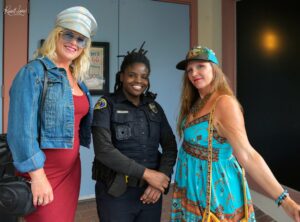 Three women smiling together: left in a denim jacket and red dress with a shiny captain's hat, center in police uniform, right in a turquoise patterned dress with a cap.