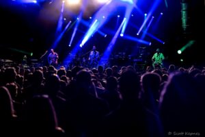 Live concert on a dark stage with blue and purple spotlights as a band performs and a crowded audience watches in the foreground.