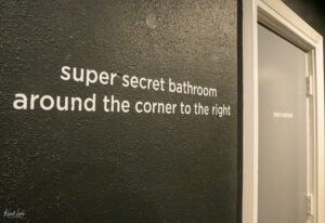 Text wall sign: dark textured wall with large white lettering reading “Super secret bathroom around the corner to the right” beside a doorway (men’s restroom visible).