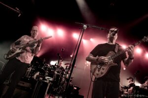 Two guitarists perform on a dark stage under red spotlights; the left musician plays a light-colored Fender-style guitar while the right guitarist plays a sunburst electric guitar, drums visible in the background.
