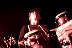 Drummer wearing a cap and glasses, illuminated by red stage light, mid-performance behind a drum kit.