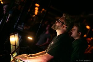 Bearded DJ wearing glasses and a cap, hands on a mixer in a dimly lit club with glowing screens and warm lights.