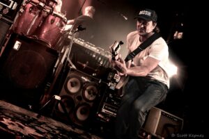 Guitarist playing an electric guitar on a dimly lit stage with amplifiers and a drummer visible in the background.