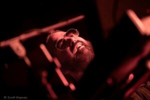 Bearded man wearing aviator sunglasses and a smile under red concert lighting, holding glow sticks in the crowd.