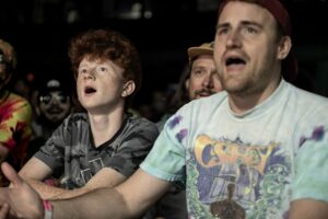 Two young men in a dark crowd with mouths open, watching a performance at night; the foreground shows a red-haired boy in a camo shirt and a man in a tie-dye shirt nearby.